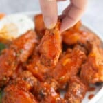 A hand holding a Buffalo chicken wing with a plate of wings behind it and a side of ranch.