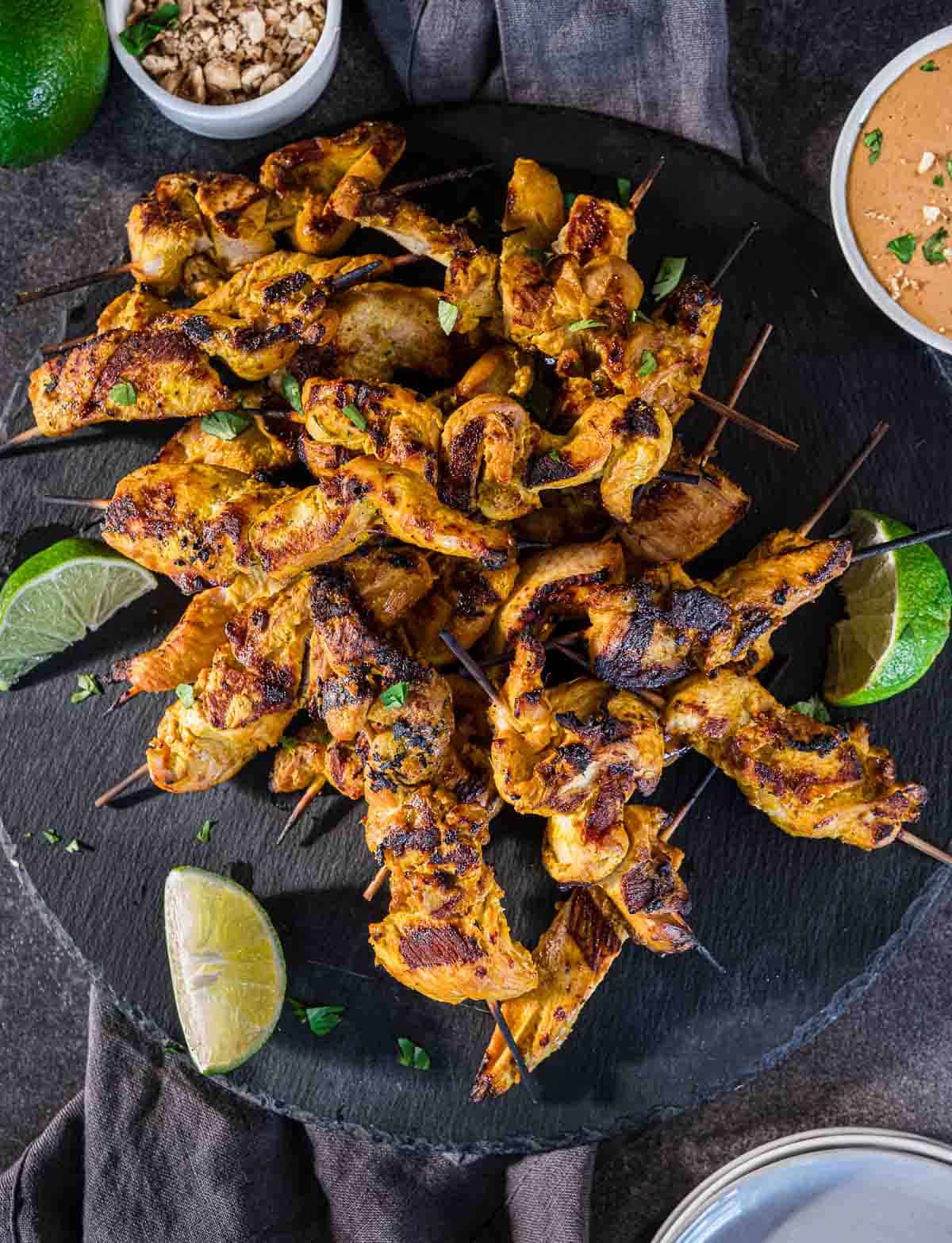 Grilled chicken satay skewers on a slate board surrounding by lime wedges.