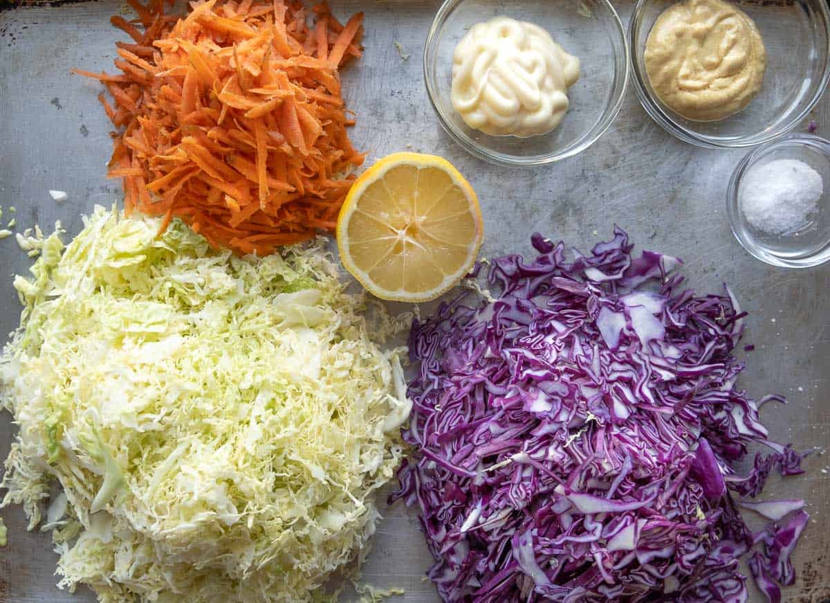 Ingredients for coleslaw including shredded carrots, green and red cabbage, mayo, Dijon mustard, lemon, and kosher salt.