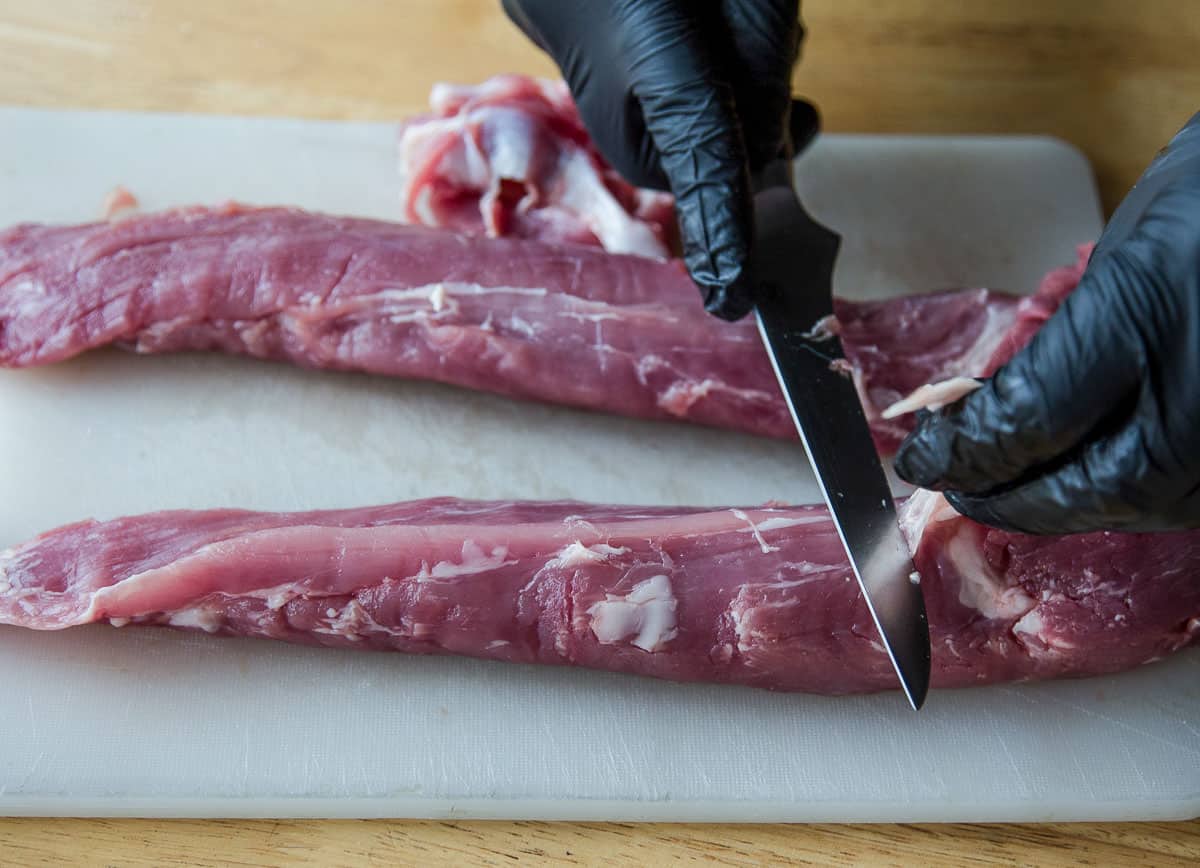 two raw pork tenderloins on a cutting board getting trimmed of excess fat