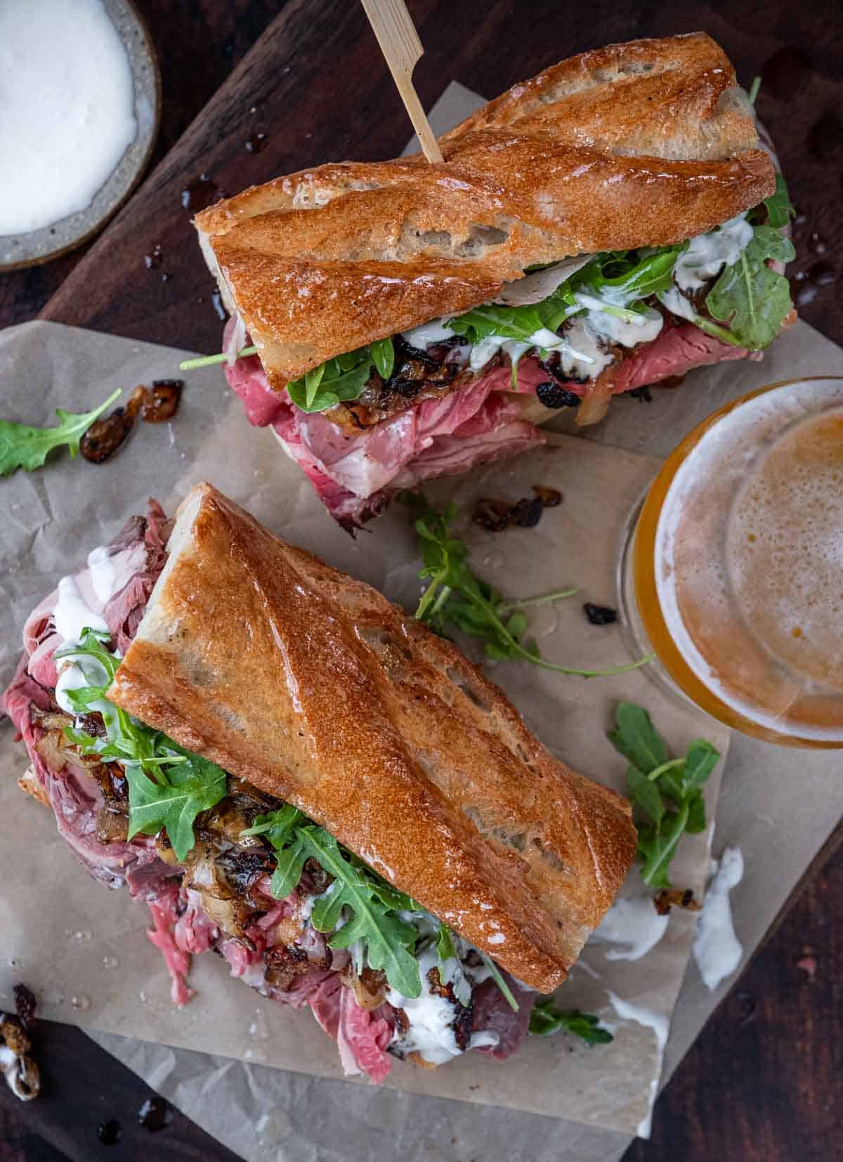 A top down photo of two prime rib steak sandwiches that are on parchment paper with a side of beer.