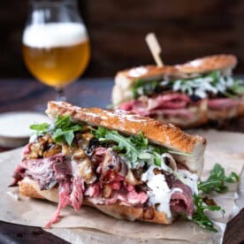 Two prime rib steak sandwiches with horseradish sauce on parchment paper on a cutting board with beer.