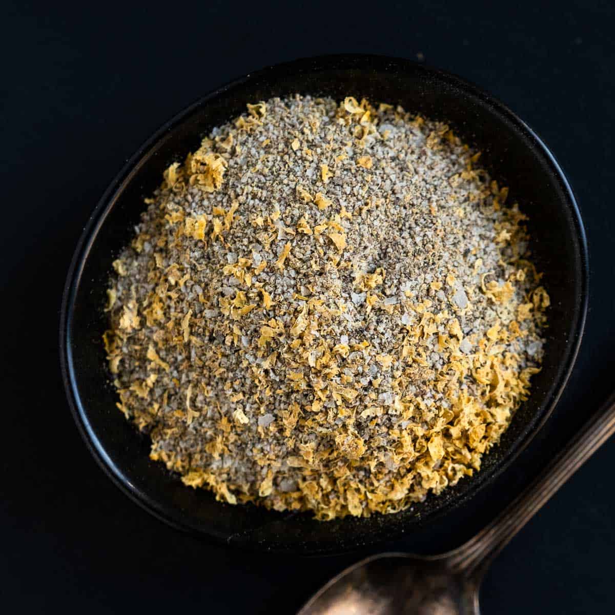 Homemade Lemon Pepper Seasoning in a black bowl with a spoon on a dark platter.