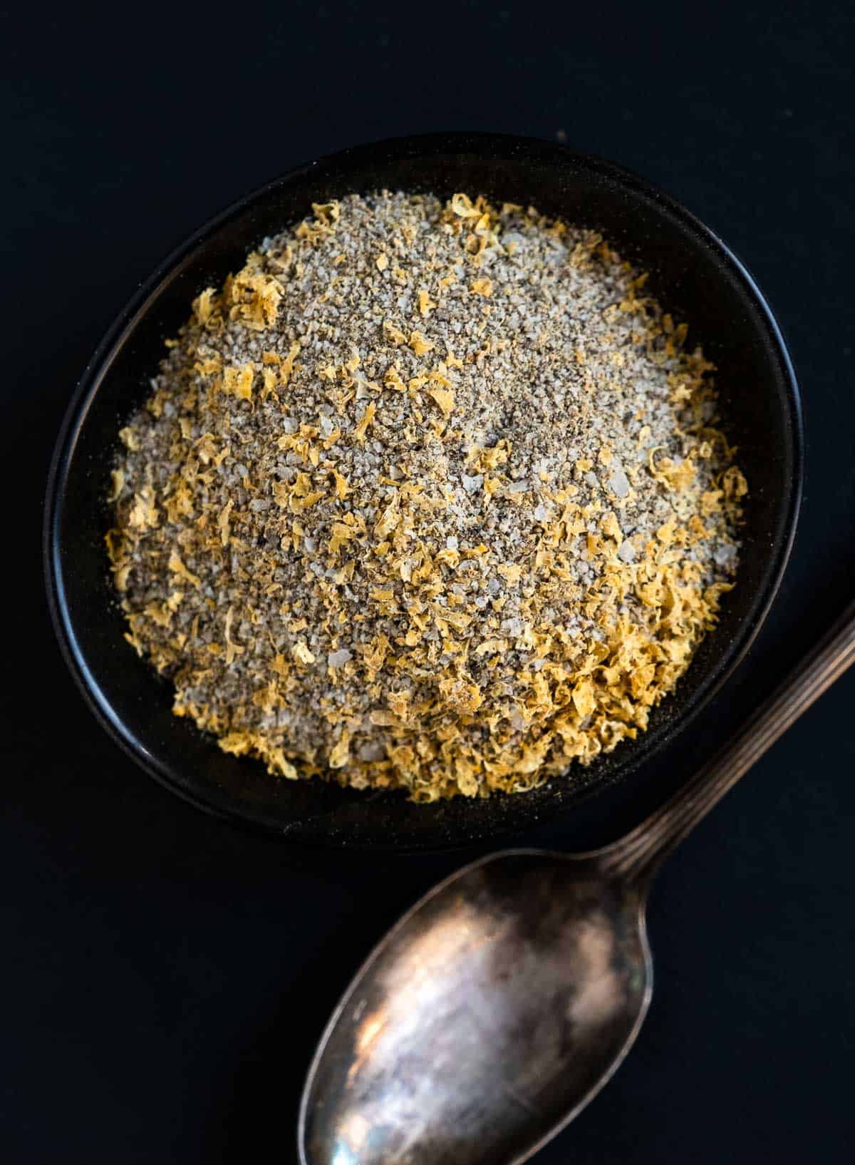 Homemade Lemon Pepper Seasoning in a black bowl with a spoon on a dark platter.