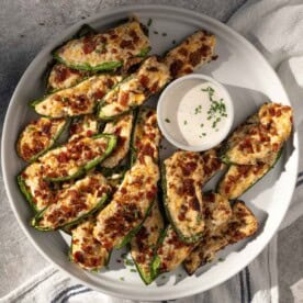 Grilled jalapeño poppers topped with crispy bacon on a serving dish with a small bowl of ranch dip.