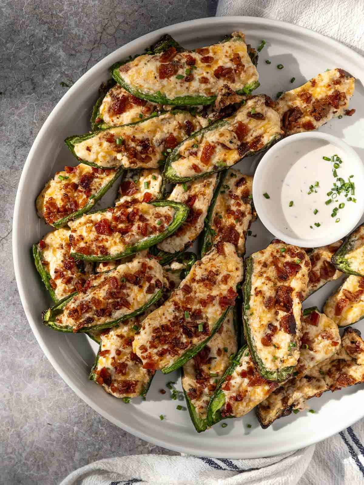 Grilled Jalapeño Poppers topped with crispy bacon on a serving platter with a small bowl of ranch dressing.