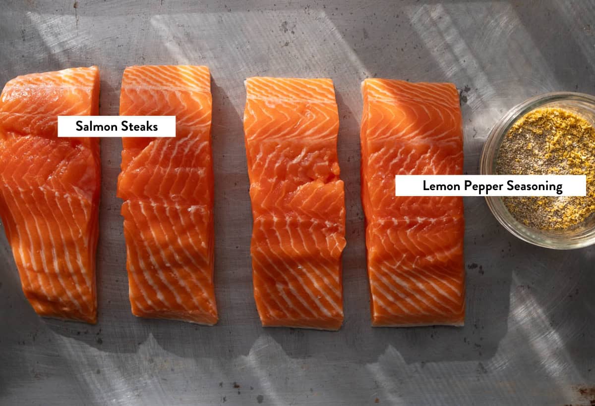 Ingredient photo of four slices of salmon and a glass bowl of lemon pepper seasoning on a baking sheet.