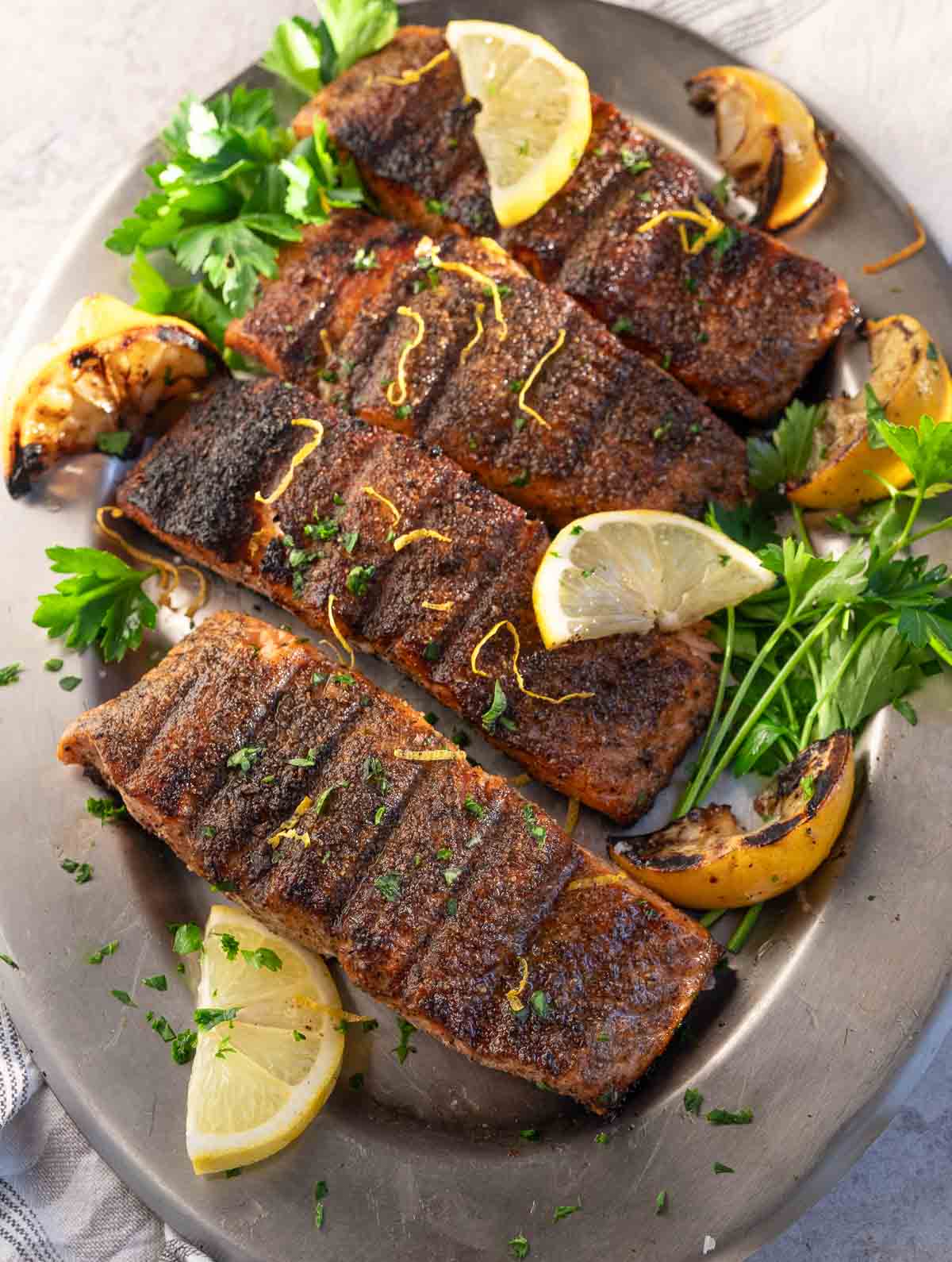 Four grilled lemon pepper salmon steaks on a silver oval platter with grilled lemon wedge and parsley garnish.