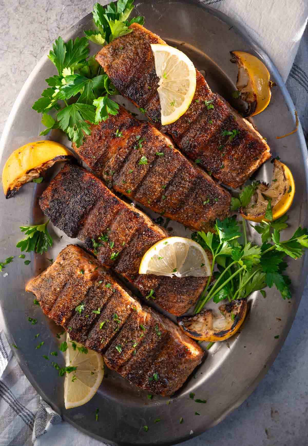 Four grilled lemon pepper salmon steaks on a silver platter with grilled lemon wedge and parsley garnish.