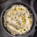 Creamy horseradish mashed potatoes in a grey bowl with a spoon topped with melted butter, chives, and cracked black pepper.