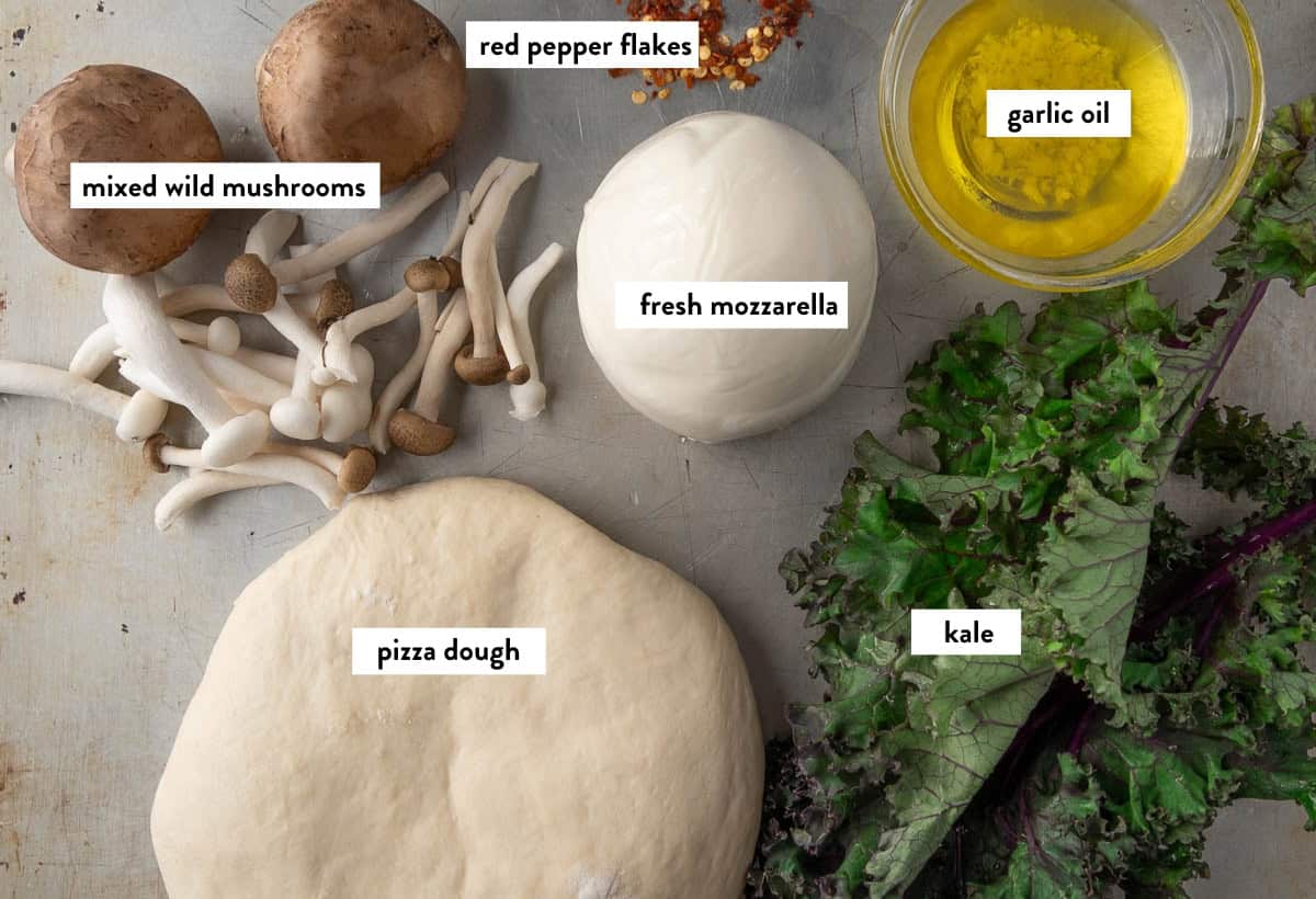 Ingredient photo for kale pizza showing dough, kale, garlic oil, mushrooms, cheese and red chili flakes on a sheet tray.