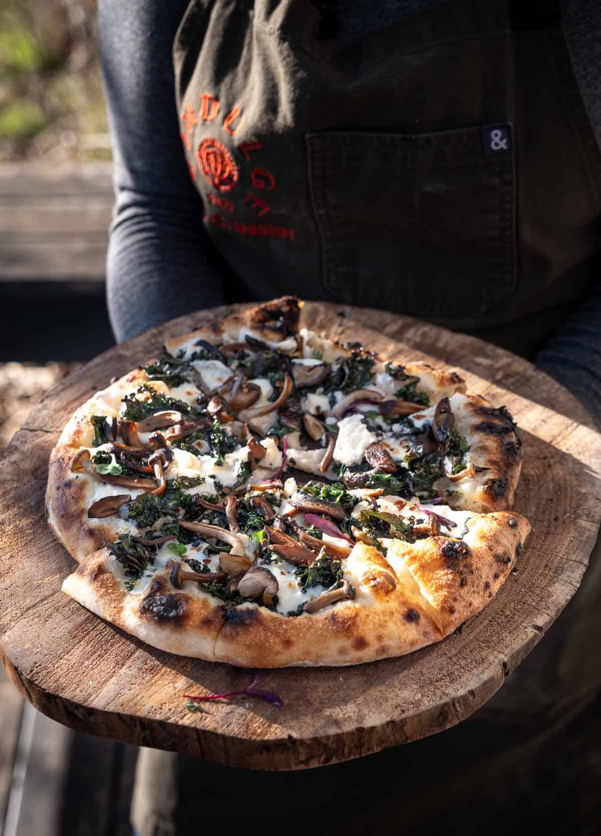 A baked kale and mushroom pizza on a wooden cutting board.